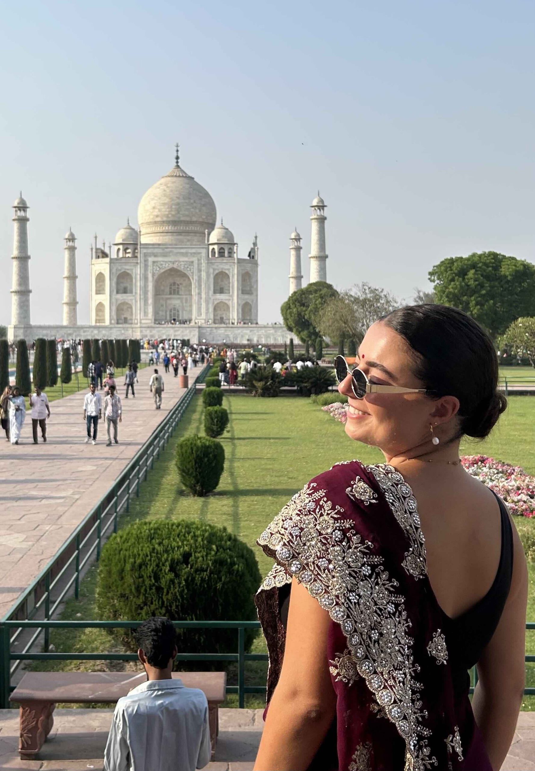 A solo female traveller standing in front of the Taj Mahal in Agra India wearing a maroon red sari and sunglasses